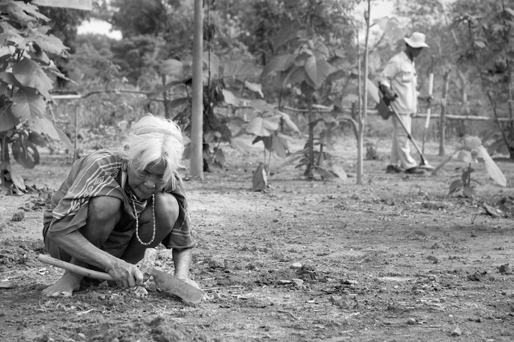 A technician with a UXO Lao bomb disposal team scans for bombs in a woman’s yard as she continues weeding. They work along a new road built atop the old Ho Chi Minh Trail. ©2006/Jerry Redfern