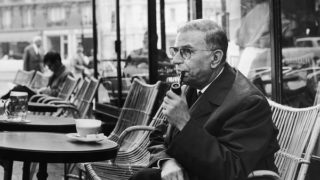 Jean-Paul Sartre smokes a pipe and drinks a coffee at a café in Paris