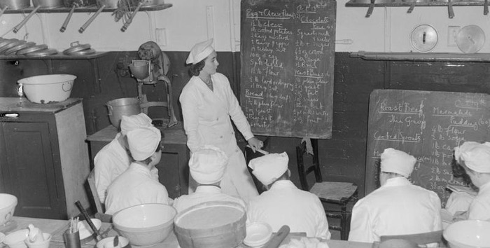 Trainee cooks during World War Two sit in front of a blackboard where a teacher is going through the recipes they are about to learn