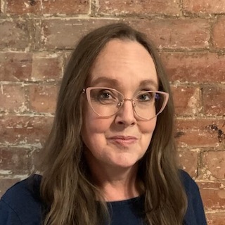 Portrait of Diane Purkiss, who has long, brown hair and large glasses and stands in front of a brick wall