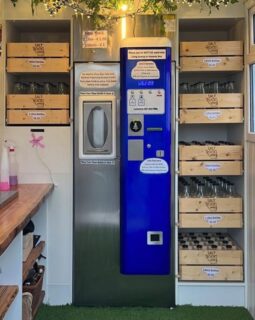 Milk vending machine. On the left is a stainless stell fronted cabinet with a recess to place bottles to receive milk. On the right is a blue panel with instructions and options for payment. Far right are racks of clean glass bottles for purchase.