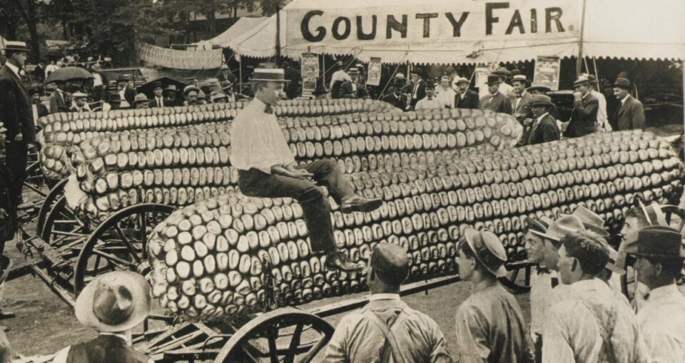 Photomontage of a man atop a giant ear of corn at a country fair with assembled onlookers gawping at the scene