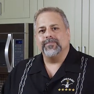 Headshot of Anthony Mongiellow, a large man with a greying Van Dyke beard, wearing a black shirt emblazoned with his company name and logo.