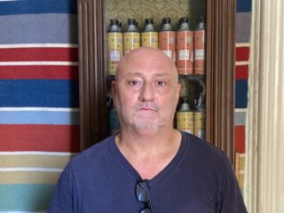 A man standing in front of a glass-fronted cupboard in which are cans of single variety olive oils.