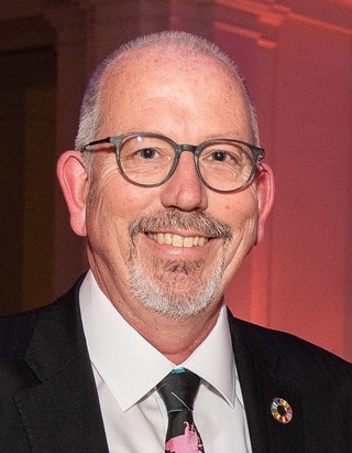 Portrait of a man with a trim beard and wearing glasses. He is smiling at the camera