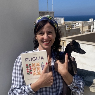 A woman with dark hair tied under a scarf holds a book and a small Italian greyhound. She is smiling at the camera and there is a blue sky and sea in the background.