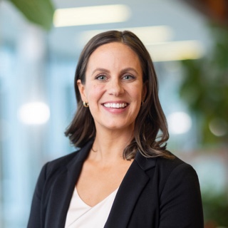 Tara Schmidt, a woman with shoulder length brown hair and blue eyes is smiling at the camera. She is wearing a dark blazer and white top. The background is blurred.
