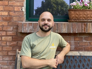 A man, balding with a beard, in a green T-shirt, sits on a bench in front of brickwork and a windowsill with flower box. Trees are reflected in the glass of the window. He is looking straight at the camera.