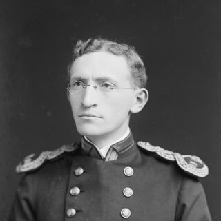 Portrait of a white man in uniform. He has wavy hair and wears spectacles.